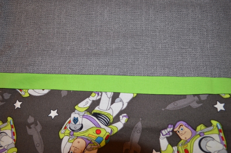 Puede incluir: Primer plano de una tela gris con un ribete verde. La tela tiene un patr&oacute;n de estrellas y naves espaciales con una imagen de dibujos animados de Buzz Lightyear.