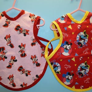May include: Two red and white bibs with a yellow trim. One bib has a pink background with a Minnie Mouse pattern. The other bib has a red background with a Minnie Mouse nautical pattern and the text "Hoo! Hoo!" repeated.