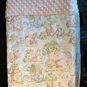 May include: A rectangular baby blanket featuring a white dimpled minky fabric top section and a bottom section printed with a colorful Winnie the Pooh pattern. The print displays various characters including Pooh, Tigger, and Eeyore in a forest setting.