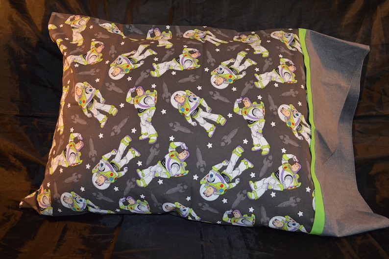 Puede incluir: Una funda de almohada gris con un patr&oacute;n repetido de Buzz Lightyear de Toy Story. La funda de almohada tiene un borde verde y gris.