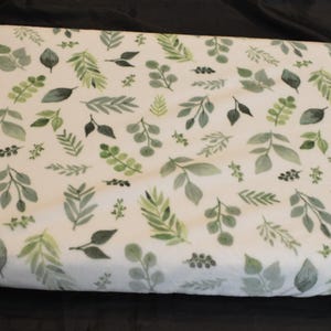 May include: A white changing pad cover with a repeating pattern of green and gray watercolor leaves. The leaves vary in size and shape, creating a natural, botanical theme. The cover is soft and likely made of a comfortable fabric.