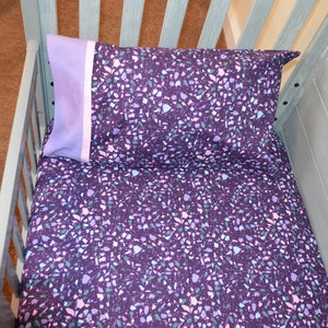 May include: A purple and white patterned crib sheet and pillow with a light purple border. The crib sheet and pillow are made of soft, comfortable fabric.