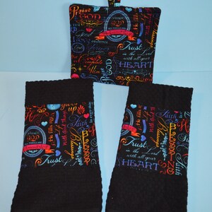 May include: Two black kitchen towels with colorful religious text printed on the bottom edge. The text includes phrases like "Trust in the Lord with all your heart" and "He fills my cup with good things". A matching pot holder with the same text is also included.