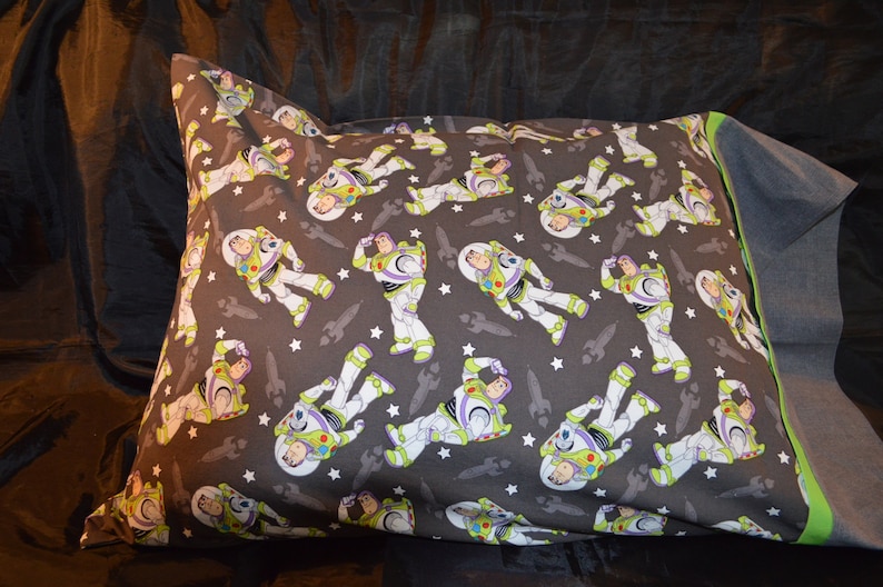Puede incluir: Una funda de almohada gris con un patr&oacute;n repetido de Buzz Lightyear de Toy Story. El patr&oacute;n incluye a Buzz Lightyear en su traje de ranger espacial, estrellas y naves espaciales. La funda de almohada tiene un borde verde.