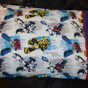 May include: A white pillowcase with a blue and red Transformers print. The pillowcase has a blue border and a red accent.