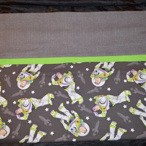 Puede incluir: Una funda de almohada de tela gris y verde con un patr&oacute;n repetido de Buzz Lightyear de Toy Story. El patr&oacute;n incluye estrellas blancas y cohetes grises.