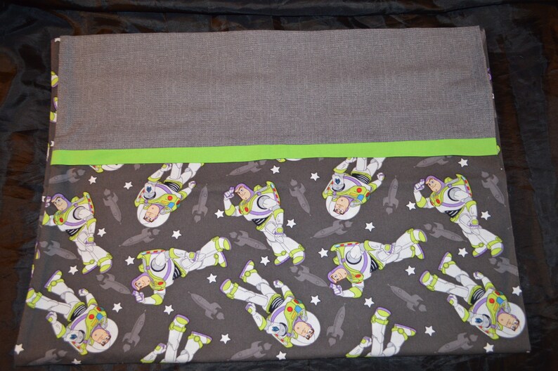 Puede incluir: Una funda de almohada de tela gris y verde con un patr&oacute;n repetido de Buzz Lightyear de Toy Story. El patr&oacute;n incluye estrellas blancas y cohetes grises.