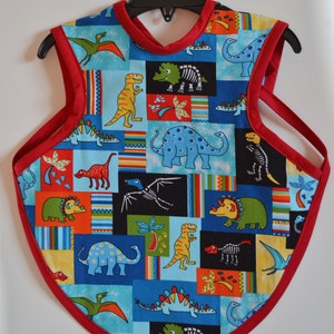 May include: A red and blue dinosaur-themed bib with a red trim. The bib has a variety of dinosaurs in different colors and patterns, including a green triceratops, a blue stegosaurus, and a yellow tyrannosaurus rex.