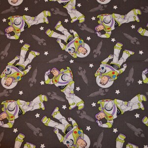 Puede incluir: Una tela gris con un patr&oacute;n repetido de un ranger espacial de dibujos animados en un traje espacial blanco y morado con una visera amarilla. El ranger espacial est&aacute; rodeado de estrellas blancas y cohetes grises.