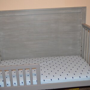 May include: A gray wooden crib with a headboard and side rails. The crib has a mattress with a blue and white pattern. The headboard has horizontal wooden planks. The crib is designed for a baby or toddler.