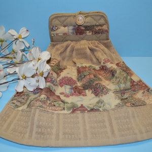 May include: A tan kitchen towel with a floral print of a cottage scene. The towel has a loop on top with a brown button. The towel is hanging on a blue background.