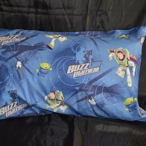 Puede incluir: Una funda de almohada azul y verde con un patrón repetido de Buzz Lightyear y personajes alienígenas de la franquicia Toy Story. La funda de almohada tiene un borde verde.