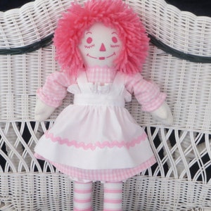 May include: A pink and white rag doll with pink yarn hair sitting on a white wicker chair.