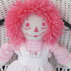 May include: A pink rag doll with a pink and white gingham dress and a white apron. The doll has pink yarn hair and embroidered facial features.
