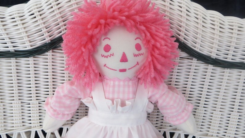 May include: A pink rag doll with a pink and white gingham dress and a white apron. The doll has pink yarn hair and embroidered facial features.