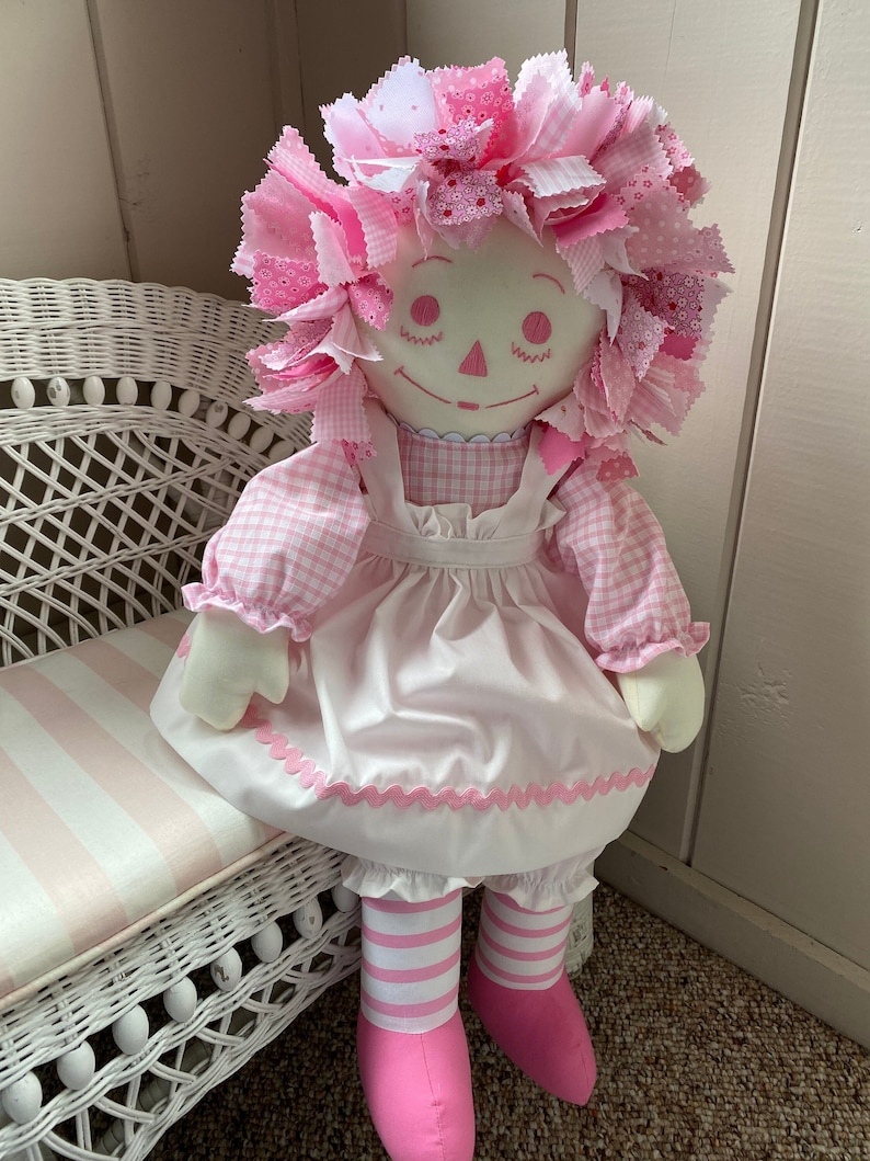 Puede incluir: Una mu&ntilde;eca de trapo rosa con un vestido de cuadros rosa y blanco, un delantal blanco con ribete rosa, medias a rayas rosa y blancas y zapatos rosas. La mu&ntilde;eca est&aacute; sentada en un banco de mimbre blanco.