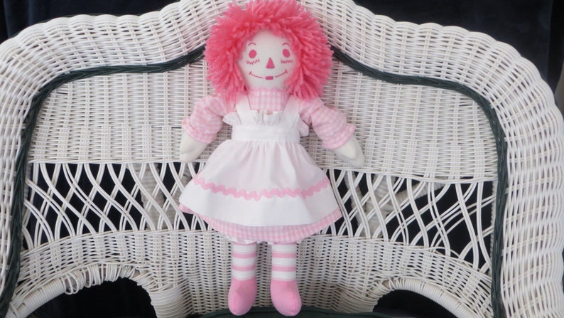 May include: A pink and white rag doll with pink yarn hair sitting on a white wicker chair.