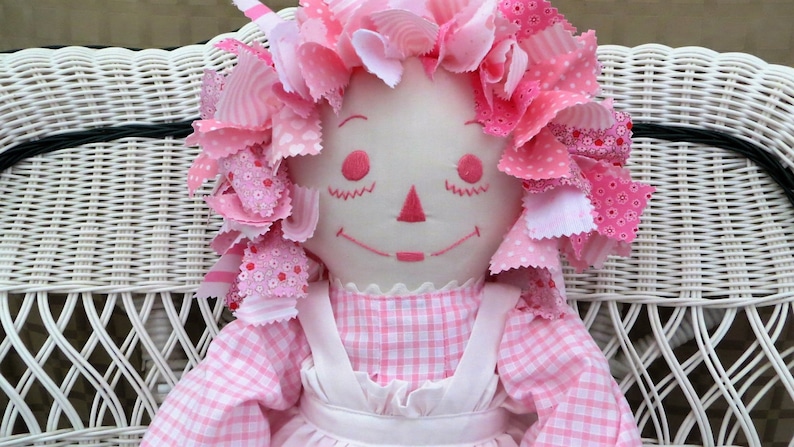 Puede incluir: Una mu&ntilde;eca de trapo rosa con la cara blanca y un vestido de cuadros rosa y blanco se sienta en una silla de mimbre blanca. La mu&ntilde;eca tiene el pelo de tela rosa con lunares blancos.
