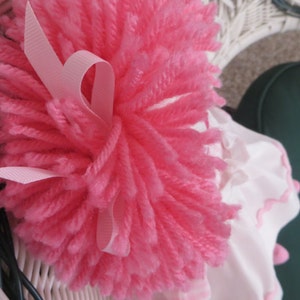 May include: A pink yarn pom-pom with a pink satin ribbon tied around it. The pom-pom is sitting on a white wicker chair.