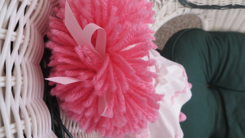 May include: A pink yarn pom-pom with a pink satin ribbon tied around it. The pom-pom is sitting on a white wicker chair.