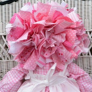 Puede incluir: Una mu&ntilde;eca de trapo rosa con un gran lazo de pelo de tela rosa y blanco. La mu&ntilde;eca lleva un vestido de cuadros rosa y blanco con un delantal blanco y un lazo blanco.