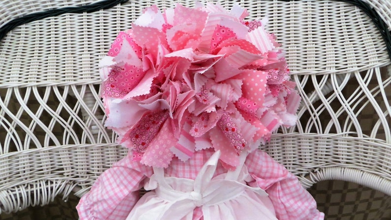 Puede incluir: Una mu&ntilde;eca de trapo rosa con un gran lazo de pelo de tela rosa y blanco. La mu&ntilde;eca lleva un vestido de cuadros rosa y blanco con un delantal blanco y un lazo blanco.