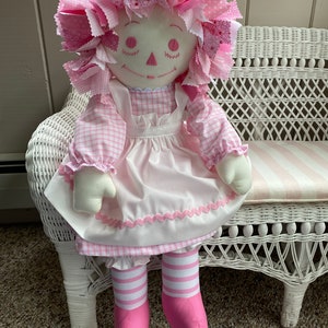Puede incluir: Una mu&ntilde;eca de trapo hecha a mano con un vestido de cuadros rosa y blanco, un delantal blanco con ribetes rosas y medias a rayas rosa y blancas. La mu&ntilde;eca tiene un lazo de pelo de tela rosa y est&aacute; sentada en un banco de mimbre blanco.