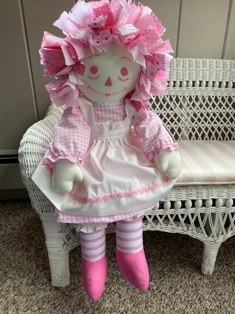 Puede incluir: Una mu&ntilde;eca de trapo hecha a mano con un vestido de cuadros rosa y blanco, un delantal blanco con ribetes rosas y medias a rayas rosa y blancas. La mu&ntilde;eca tiene un lazo de pelo de tela rosa y est&aacute; sentada en un banco de mimbre blanco.