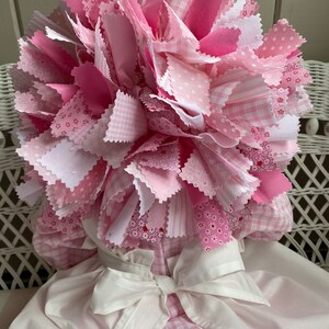 Puede incluir: Una mu&ntilde;eca de tela rosa y blanca con una gran flor de tela rosa en la cabeza. La mu&ntilde;eca lleva un vestido blanco con una falda de cuadros rosa y blanca.