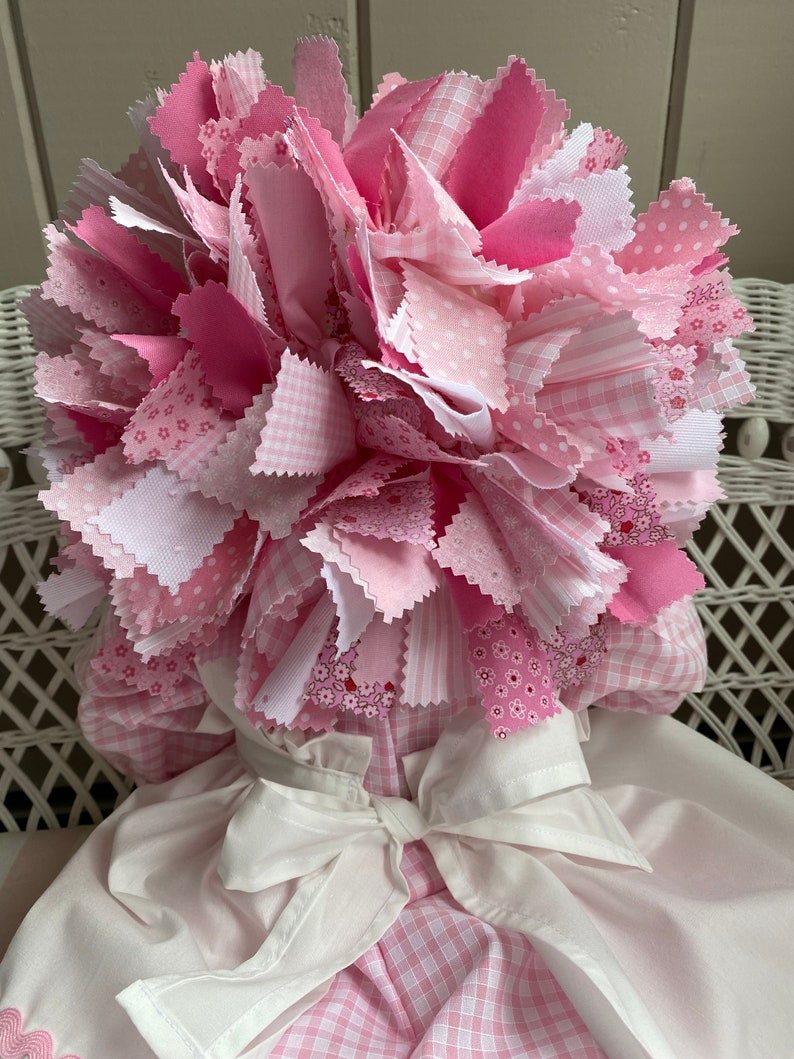 Puede incluir: Una mu&ntilde;eca de tela rosa y blanca con una gran flor de tela rosa en la cabeza. La mu&ntilde;eca lleva un vestido blanco con una falda de cuadros rosa y blanca.