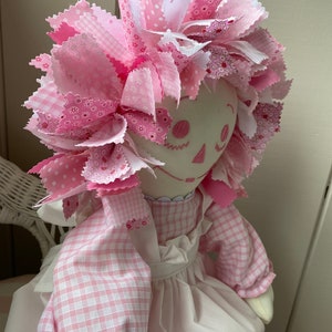Puede incluir: Una mu&ntilde;eca de trapo rosa con un vestido blanco y mangas de cuadros rosas. La mu&ntilde;eca tiene el pelo de tela rosa y blanco, una cara blanca y ojos y boca bordados.