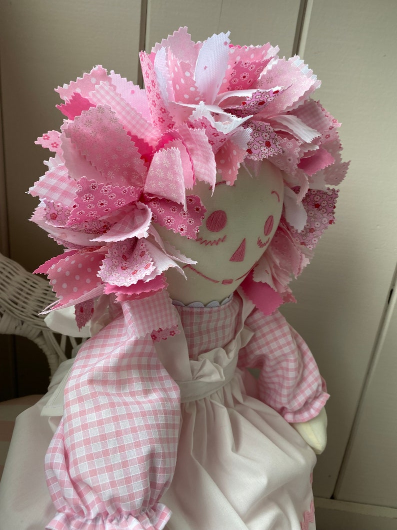 Puede incluir: Una mu&ntilde;eca de trapo rosa con un vestido blanco y mangas de cuadros rosas. La mu&ntilde;eca tiene el pelo de tela rosa y blanco, una cara blanca y ojos y boca bordados.