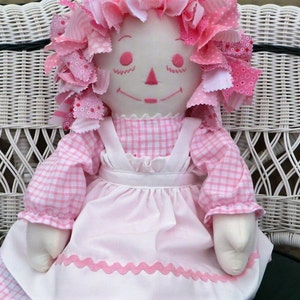 Puede incluir: Una mu&ntilde;eca de trapo rosa con un vestido de cuadros y un delantal blanco se sienta en una silla de mimbre blanca. La mu&ntilde;eca tiene el pelo de tela rosa y blanco y una cara sonriente.