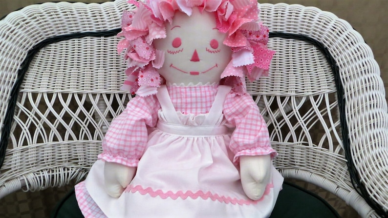 Puede incluir: Una mu&ntilde;eca de trapo rosa con un vestido de cuadros y un delantal blanco se sienta en una silla de mimbre blanca. La mu&ntilde;eca tiene el pelo de tela rosa y blanco y una cara sonriente.