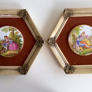 May include: Two framed decorative porcelain plaques. Each plaque features a round, hand-painted scene of a couple in a garden setting. The frames are hexagonal with ornate gold accents and a burgundy background.