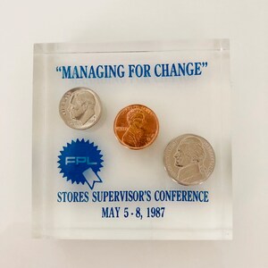 May include: A clear acrylic plaque with the text "Managing for Change" and a blue starburst with the letters "FPL" inside. There are three coins inside the plaque: a silver dime, a copper penny, and a silver half dollar. The text "Stores Supervisor's Conference May 5-8, 1987" is printed below the coins.