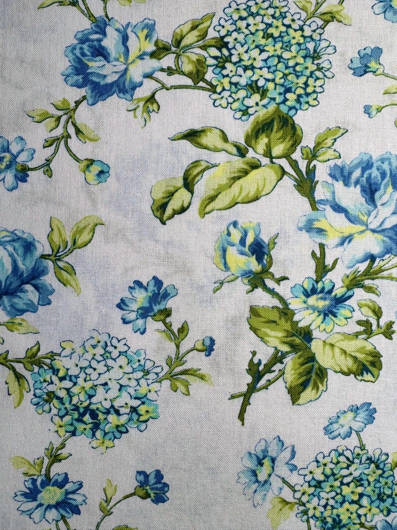 K&ouml;nnte beinhalten: Blumenstoff mit blauen und wei&szlig;en Blumen, gr&uuml;nen Bl&auml;ttern und Stielen. Das Design umfasst gro&szlig;e Rosen und kleinere Bl&uuml;tencluster, die ein Muster auf wei&szlig;em Hintergrund bilden.