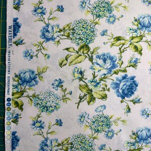 K&ouml;nnte beinhalten: Wei&szlig;er Stoff mit floralem Muster mit blauen Rosen und Hortensienbl&uuml;ten, akzentuiert durch gr&uuml;ne Bl&auml;tter. Der Stoff ist mit dem Text "WAVERLY INSPIRATIONS" bedruckt.