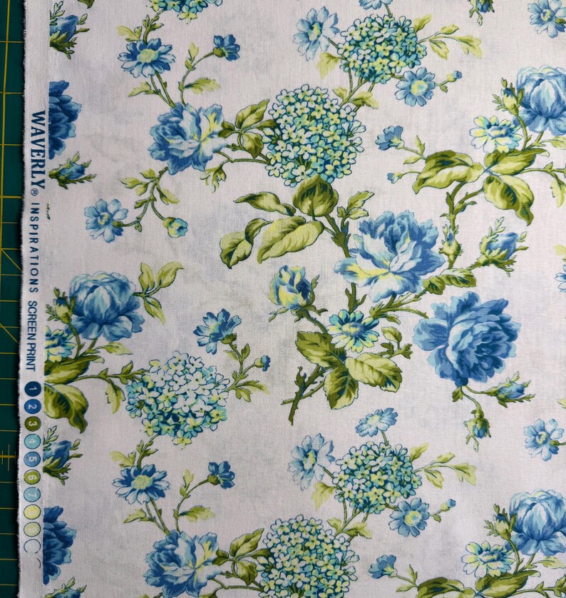K&ouml;nnte beinhalten: Wei&szlig;er Stoff mit floralem Muster mit blauen Rosen und Hortensienbl&uuml;ten, akzentuiert durch gr&uuml;ne Bl&auml;tter. Der Stoff ist mit dem Text "WAVERLY INSPIRATIONS" bedruckt.