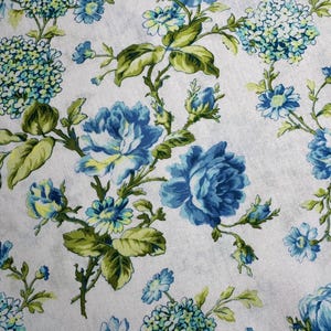 K&ouml;nnte beinhalten: Nahaufnahme eines wei&szlig;en Stoffes mit floralem Muster. Das Design zeigt gro&szlig;e blaue Rosen, kleinere blaue und gelbe Bl&uuml;ten sowie gr&uuml;ne Bl&auml;tter und Stiele. Die Gesamt&auml;sthetik ist vintage und botanisch.