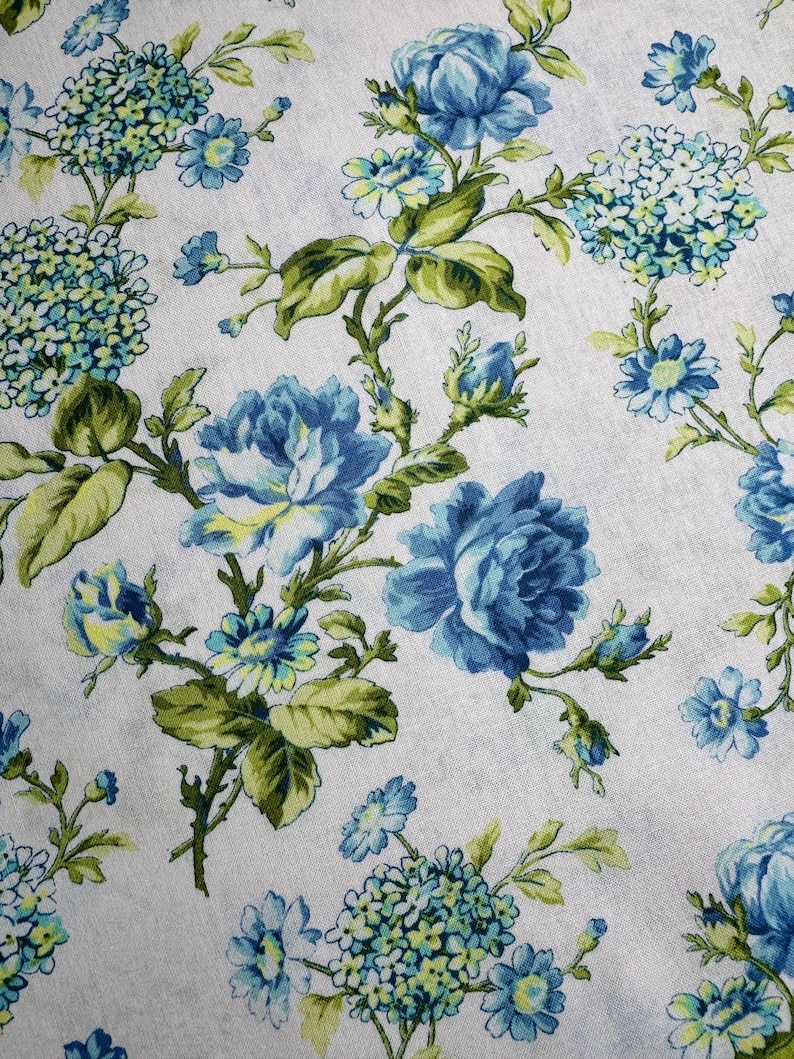 K&ouml;nnte beinhalten: Nahaufnahme eines wei&szlig;en Stoffes mit floralem Muster. Das Design zeigt gro&szlig;e blaue Rosen, kleinere blaue und gelbe Bl&uuml;ten sowie gr&uuml;ne Bl&auml;tter und Stiele. Die Gesamt&auml;sthetik ist vintage und botanisch.
