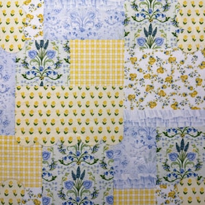 May include: A patchwork textile featuring a variety of patterns. The design incorporates squares of yellow gingham, floral prints with yellow and blue flowers, and blue damask patterns. Some squares have ruffled edges, creating a textured appearance.