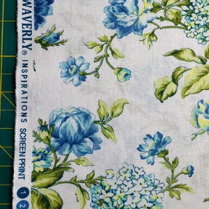 K&ouml;nnte beinhalten: Wei&szlig;er Stoff mit floralem Muster. Gro&szlig;e blaue und hellblaue Blumen, gr&uuml;ne Bl&auml;tter und der Text "WAVERLY INSPIRATIONS SCREEN PRINT". Der Stoff hat eine Vintage-&Auml;sthetik.