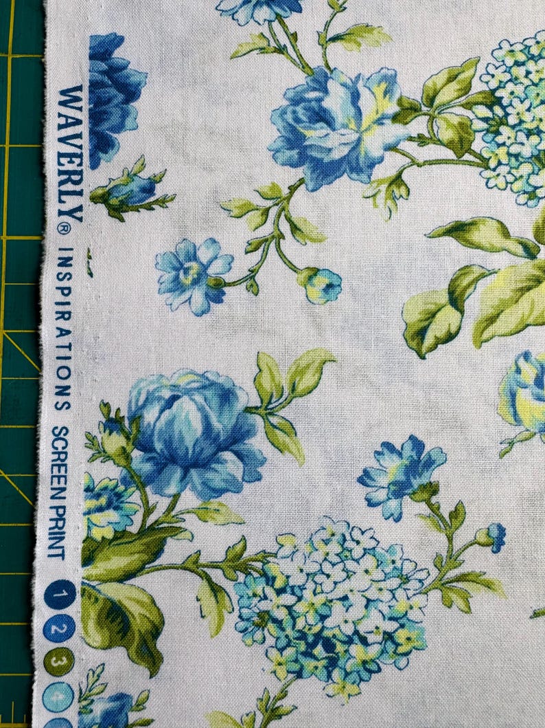 K&ouml;nnte beinhalten: Wei&szlig;er Stoff mit floralem Muster. Gro&szlig;e blaue und hellblaue Blumen, gr&uuml;ne Bl&auml;tter und der Text "WAVERLY INSPIRATIONS SCREEN PRINT". Der Stoff hat eine Vintage-&Auml;sthetik.