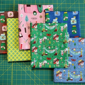 May include: A collection of folded fabric pieces with various patterns. Designs include insects in jars, camping scenes, and whimsical forest motifs with mushrooms and animals. Colors range from brown and green to pink and blue.