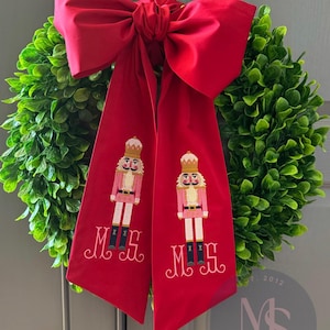 May include: A green boxwood wreath with a large red bow and two red ribbons. Each ribbon features an embroidered nutcracker soldier and the initials "MS". The wreath is a festive holiday decoration.