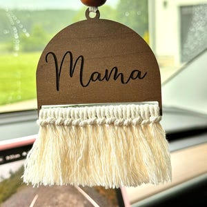 May include: Wooden car charm with the word "Mama" engraved on it. The charm has a fringe of white yarn and is hanging from a string with two wooden beads.