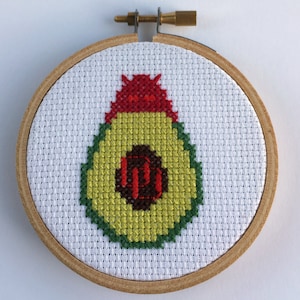 Puede incluir: Bordado de punto de cruz de un aguacate, con piel verde, pulpa amarilla y hueso marrón. El aguacate está centrado en un bastidor de bordado de madera. La parte superior del aguacate es roja. El fondo es blanco.