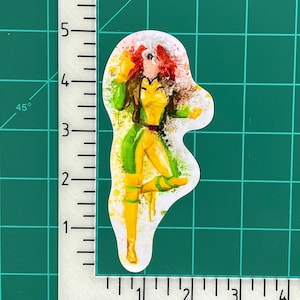 May include: A hand-painted sticker featuring a superhero character with red hair, a yellow and green costume, and a white background. The sticker is approximately 6 inches tall.