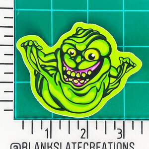 May include: Green and yellow cartoon ghost with a wide, toothy grin and pink tongue. The ghost has a large, round body and small, beady eyes. The ghost is drawn in a cartoon style and has a mischievous expression.